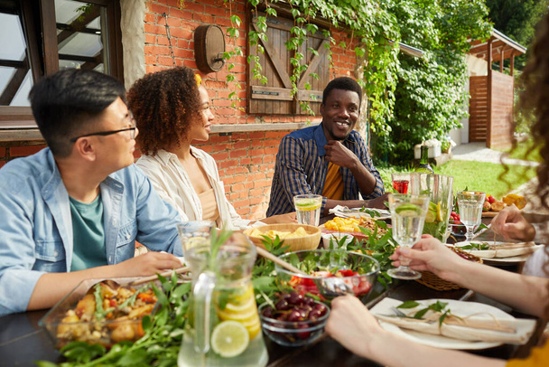 オープンテラスのテーブルに座っている間に屋外で一緒に夕食を楽しむ友人の多民族グループの肖像画、アフリカ系アメリカ人の男性の共有の物語、コピースペースに焦点を当てる - 写真・画像