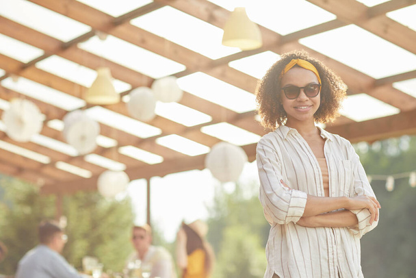 Portrait taille haute de jeune femme afro-américaine souriant à la caméra au soleil, tout en posant à l'extérieur en été avec des amis et de la famille appréciant le fond du dîner, l'espace de copie - Photo, image
