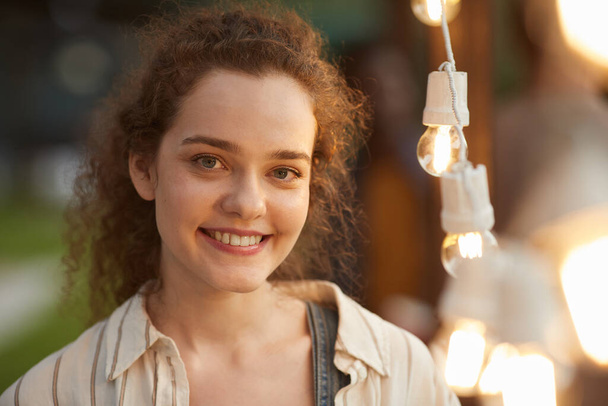 Retrato franco de cerca de una joven sonriente mirando a la cámara mientras está de pie junto a las luces en la fiesta al aire libre - Foto, Imagen
