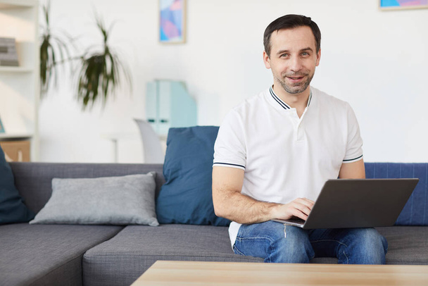 Portrait of smiling mature man using laptop and looking at camera while sitting on couch at home or in office, copy space - Foto, Imagem