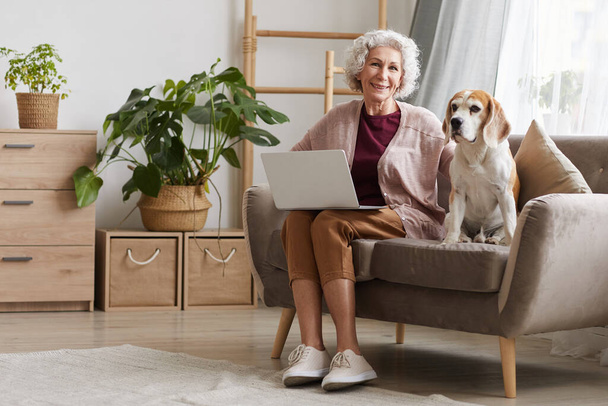 Full length portrait of modern senior woman using laptop while sitting with dog on sofa in cozy apartment and smiling at camera, copy space - Foto, Bild