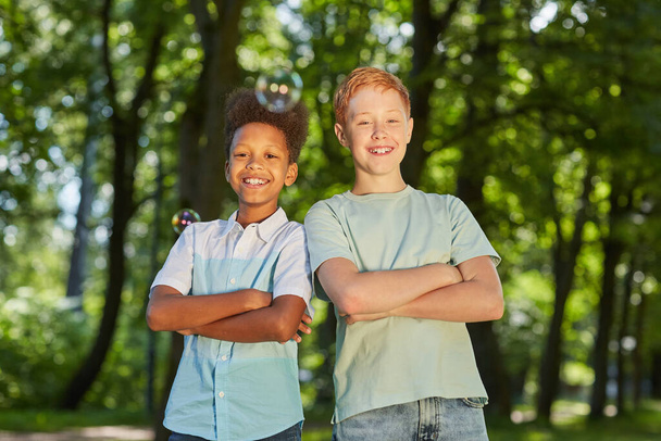 Obraz dvou usmívajících se dospívajících chlapců stojících spolu v parku a dívajících se na kameru s mýdlovými bublinami v popředí, kopírovací prostor - Fotografie, Obrázek
