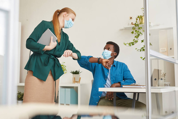 Portrait latéral du jeune homme afro-américain portant un masque facial cognant les coudes avec une collègue pendant qu'il travaillait dans une cabine au bureau post-pandémique, espace de copie - Photo, image