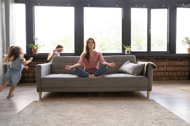 Calm woman concentrating on yoga exercises on couch at home while two noisy kids laughing, running - Foto, afbeelding