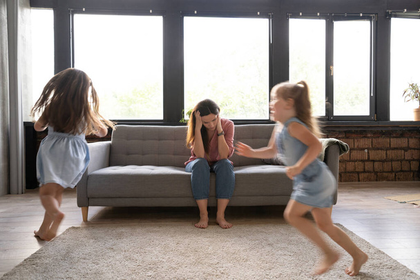Young tired mother suffers from headache closed eyes touch forehead sitting on couch while her daughters running around - Foto, Imagen
