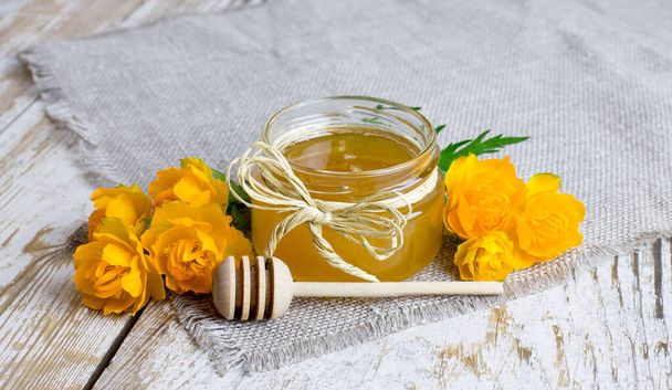 een kleine glazen pot honing met een houten lepel en trol bloemen close-up op een houten achtergrond in een rustieke stijl. - Foto, afbeelding