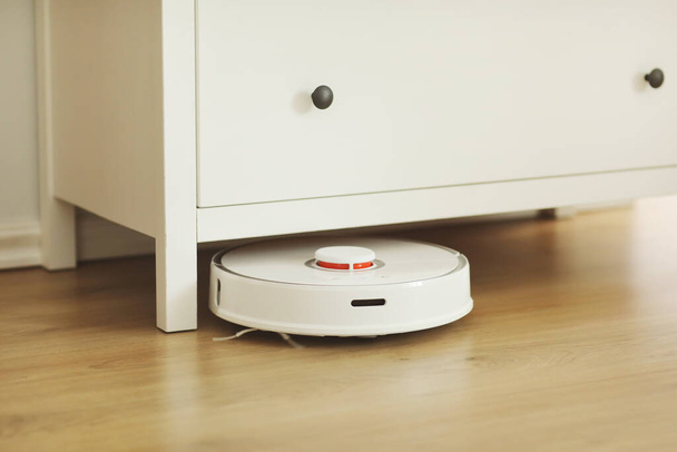 Witte robot stofzuiger op laminaat vloer reinigen stof in woonkamer interieur. Slimme elektronische huishoudtechnologie. slimme woning. - Foto, afbeelding