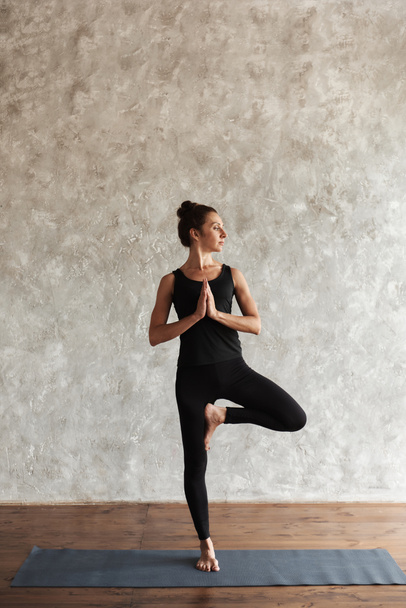 Strong middle-aged woman practicing yoga, looking away concentrated, doing Vrksasana exercise, Tree pose, working out, wearing black sportswear at yoga studio with gray wall. Healthy life concept. - Φωτογραφία, εικόνα