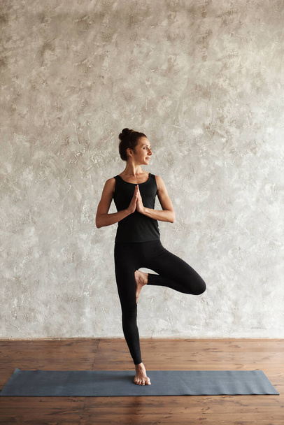Yogi mid aged smiling woman practicing yoga, looking away concentrated, doing Vrksasana exercise, Tree pose, working out, wearing black sportswear at home with gray wall. Healthy life concept. - 写真・画像