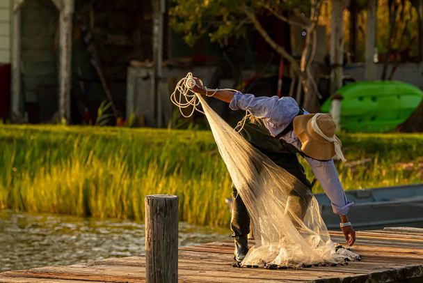 A fisherman wearing overalls, plastic boots and a bucket hat is preparing his cast net before throwing it into the Chesapeake Bay at sunset. He is on a wooden pier and sun reflects from water surface. - Photo, Image