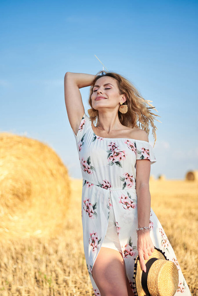 Молодая блондинка, одетая в длинное белое романтическое платье, держит соломенную шляпу и сушеный травяной букет, позирует перед пшеничным тюком на поле летом. Женский портрет на естественном фоне. - Фото, изображение