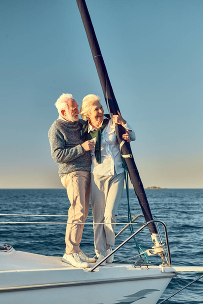 Full leghth of beautiful happy senior couple in love, retired man and woman standing on the side of sail boat floating in sea, hugging and enjoying view - Photo, Image