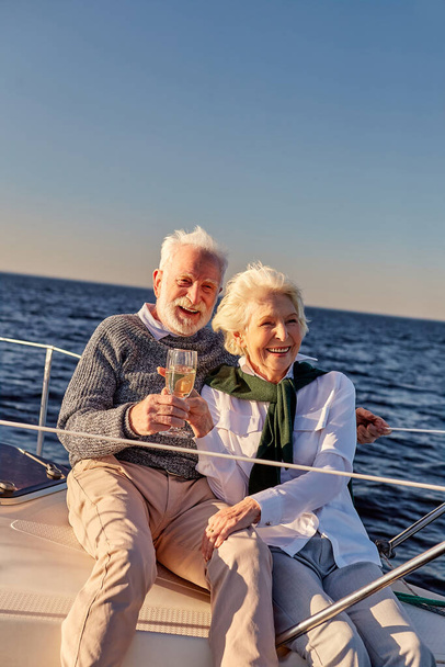 Εις υγείαν Ευτυχισμένο ζευγάρι ηλικιωμένων πίνοντας κρασί ή σαμπάνια και χαμογελώντας γιορτάζοντας την επέτειο του γάμου σε ένα ιστιοφόρο ή κατάστρωμα γιοτ επιπλέουν σε μια ήρεμη γαλάζια θάλασσα - Φωτογραφία, εικόνα