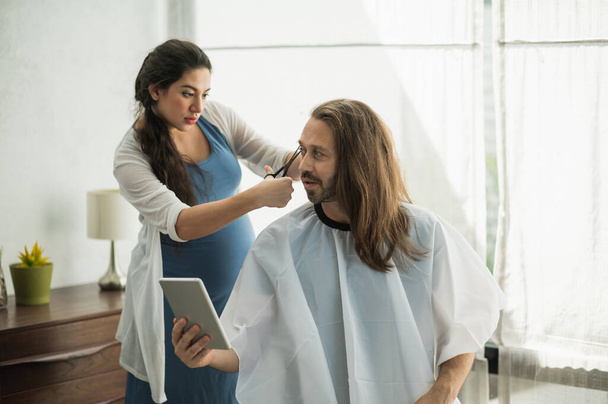 The pregnant woman is cutting her husband. Bearded man getting haircut by his wife at home new normal concept. - Фото, изображение
