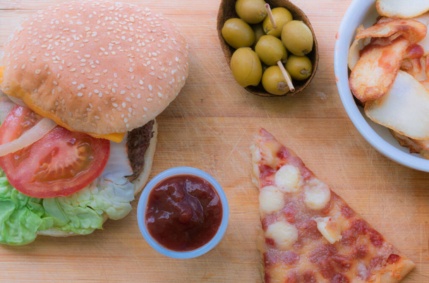 Kousek pizzy Margherita, olivy, hamburger a hranolky na dřevěném stole. Koncept pojídání odpadků. Horní pohled. - Fotografie, Obrázek