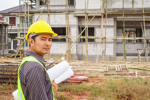 Ασιάτης επιχειρηματίας μηχανικός κατασκευών εργαζόμενος σε προστατευτικό κράνος και σχεδιαγράμματα χαρτί στο χέρι στο σπίτι εργοτάξιο - Φωτογραφία, εικόνα