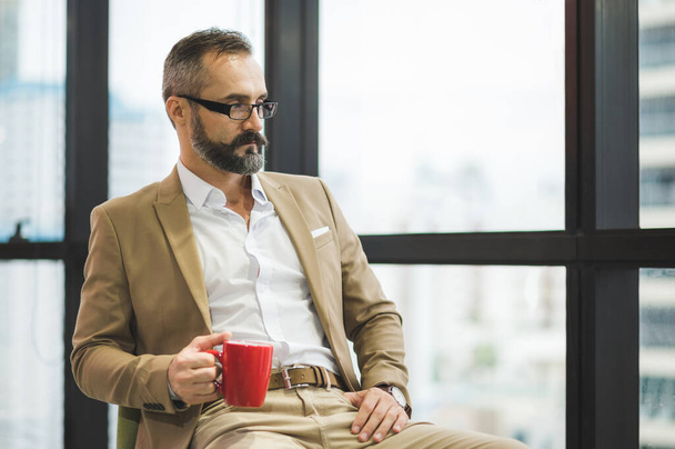Young handsome business bearded man drinking cup of coffee sitting near window in office work place. - Foto, Imagem