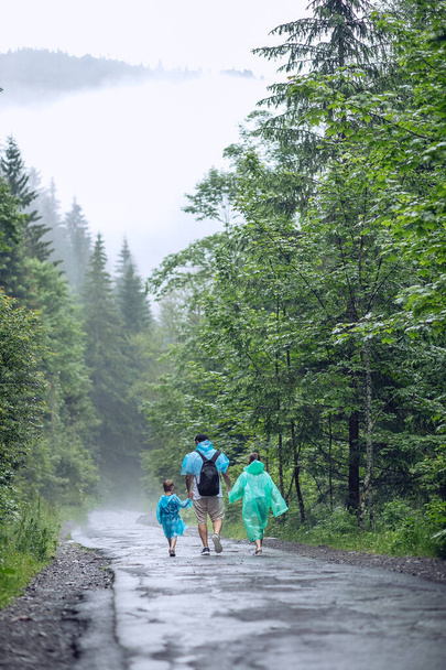 Mountain landscape. Father, son and daughter are walking on a wet road after a heavy rain in raincoats. Children hold their father's hand - Фото, изображение