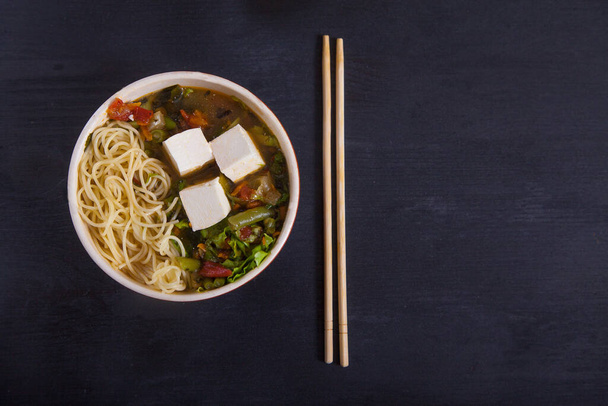 Japanese soup with tofu, noodles, vegetables and herbs, bamboo chopsticks on a black background. Copy spaes - Photo, Image