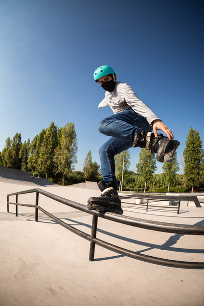 male roller skater with jeans and t-shirt, protective helmet and coronavirus prevention mask (new post covid normality) does a trick in a skate park by toasting on a railing. - Fotografie, Obrázek
