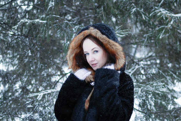 Young smiling beautiful woman with blue eyes in black faux fur coat with red hood on background of winter snowy park. - Fotografie, Obrázek