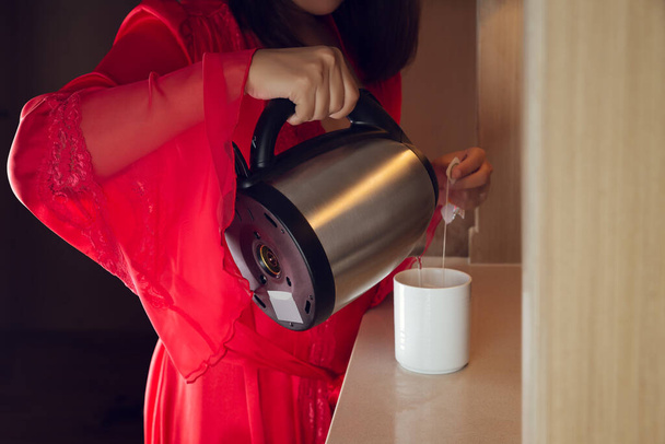 Naisella oli punainen satiininpitkä kylpytakki keittämässä teetä keittiössä öisin. Aasian tyttö kaada kuumaa vettä valkoinen kuppi - Valokuva, kuva
