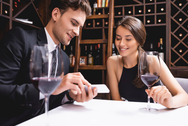 Selective focus of man in suit showing smartphone to girlfriend with glass of wine in restaurant  - Foto, Imagen