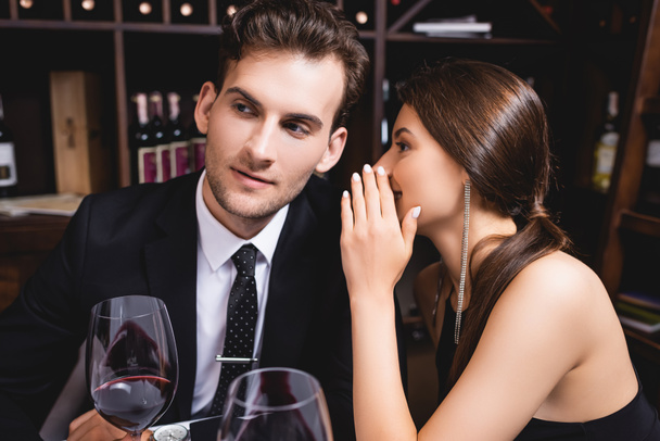 Selective focus of woman whispering to boyfriend in suit near glasses of wine in restaurant  - 写真・画像