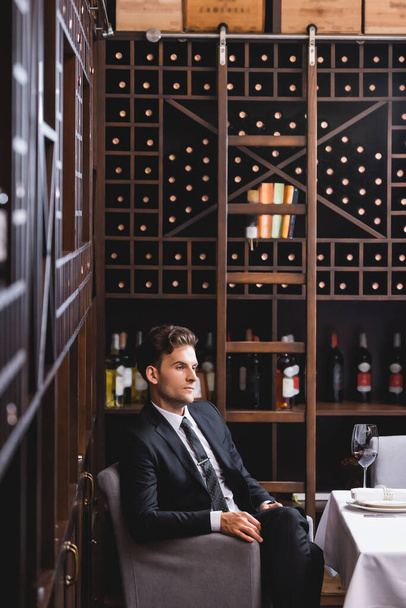 Selektiver Fokus eines jungen Mannes im Anzug, der in der Nähe von Weinglas und Regalen mit Weinflaschen im Restaurant sitzt  - Foto, Bild