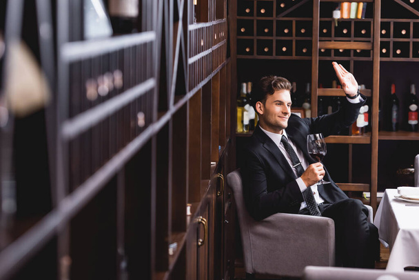 Selective focus of young man in suit waving hand and holding glass of wine in restaurant  - 写真・画像