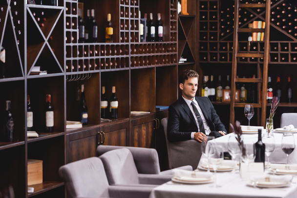 Concentration sélective de l'homme en tenue de cérémonie assis près du verre de vin et racks avec des bouteilles dans le restaurant  - Photo, image
