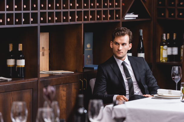 Concentration sélective du jeune homme en costume regardant loin tout en étant assis près d'un verre de vin sur la table dans le restaurant  - Photo, image