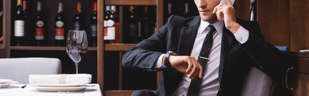 Panoramic shot of man in suit talking on smartphone and checking time on wristwatch near glass of wine in restaurant  - Photo, Image