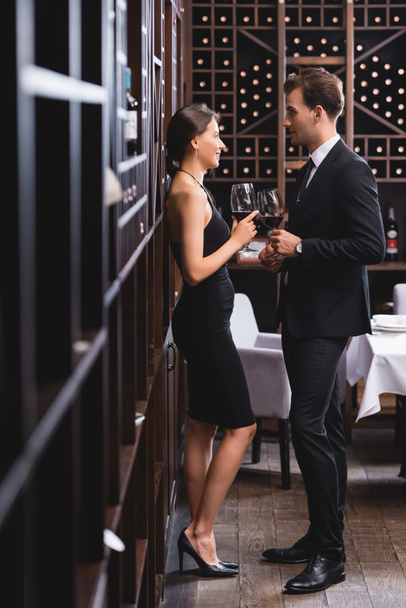 Selective focus of man in suit holding glass of wine during dating with elegant girlfriend in restaurant  - Foto, imagen