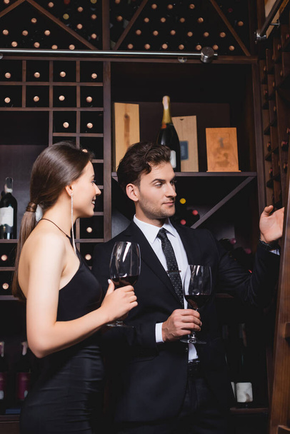 Man in suit pointing with hand at rack with bottles near elegant woman with glass of wine in restaurant  - Φωτογραφία, εικόνα
