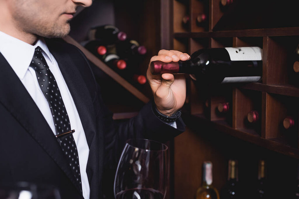 Ausgeschnittene Ansicht eines Mannes im Anzug, der eine Flasche aus einem Regal in der Nähe eines Weinglases in einem Restaurant nimmt  - Foto, Bild