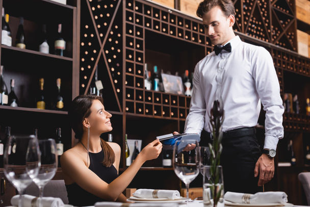 Selective focus of young woman holding credit card near waiter in formal wear with payment terminal in restaurant  - Photo, Image