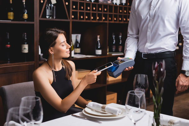 Enfoque selectivo de mujer elegante pagando con smartphone a camarero en restaurante  - Foto, imagen