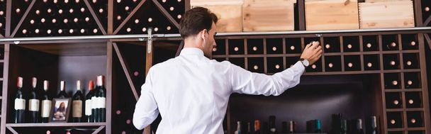 Панорамный снимок сомелье, берущего винную бутылку со стойки в ресторане  - Фото, изображение