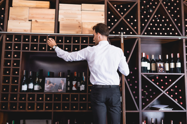 Rückansicht eines Sommeliers in formeller Kleidung, der in einem Restaurant eine Flasche Wein aus dem Regal holt  - Foto, Bild