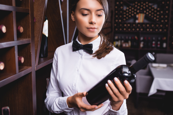 Concentration sélective du jeune sommelier en tenue formelle regardant la bouteille de vin dans le restaurant  - Photo, image