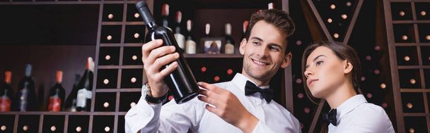 Ιστοσελίδα κεφαλίδα του sommelier κρατώντας μπουκάλι κρασί κοντά στο συνάδελφο στο εστιατόριο  - Φωτογραφία, εικόνα