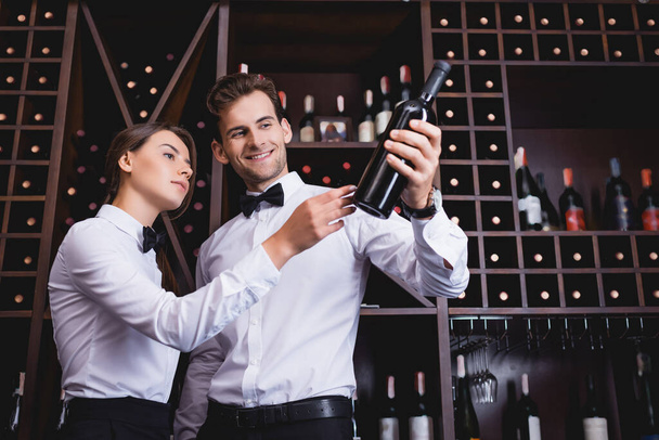 Young sommelier holding bottle of wine near colleague and racks in restaurant  - Foto, afbeelding