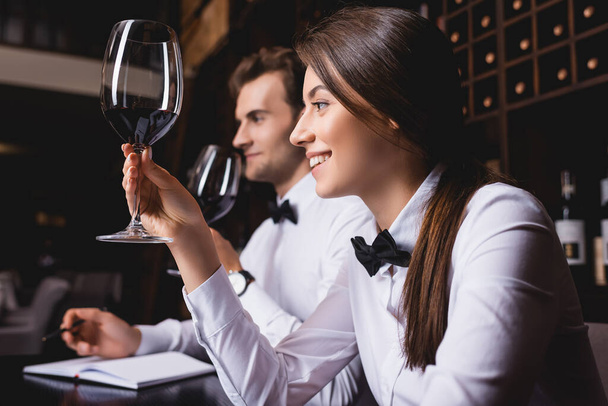 Focus selettivo del giovane sommelier che tiene un bicchiere di vino vicino al collega nel ristorante  - Foto, immagini