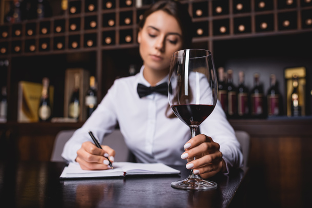 Focus selettivo del sommelier che tiene un bicchiere di vino mentre scrive sul taccuino a tavola  - Foto, immagini