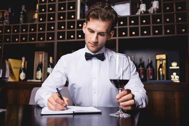 Selective focus of sommelier in formal wear writing on notebook during degustation of wine  - Foto, Imagen