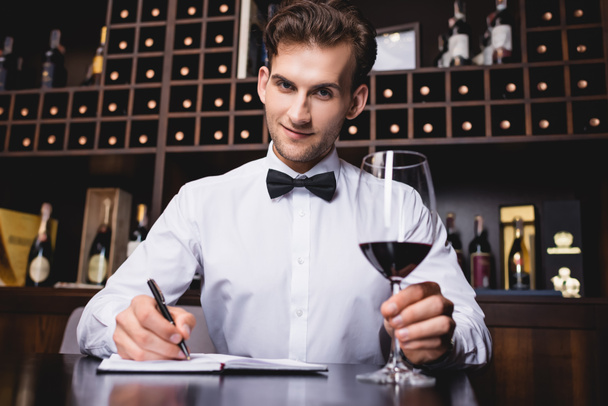 Селективный фокус сомелье смотрит в камеру, держа бокал вина и записывая на ноутбук в ресторане  - Фото, изображение