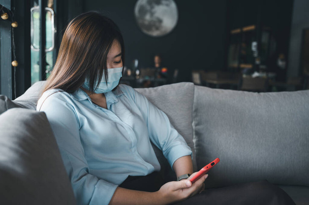 Asian woman wearing a mask uses a smartphone in a coffee shop. - Zdjęcie, obraz