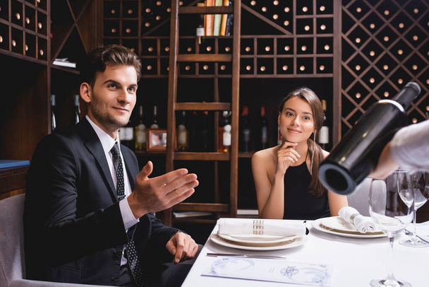 Selective focus of man pointing at sommelier with bottle of wine near girlfriend in restaurant  - Фото, изображение