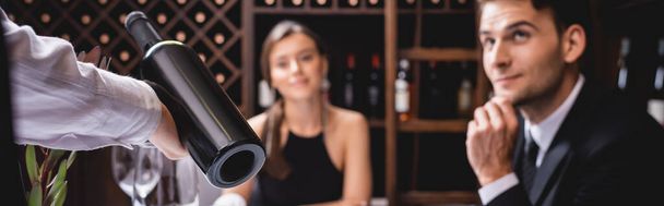 Sommelier 'in yatay mahsulü restoranda zarif bir çiftin yanında bir şişe şarap tutuyor.  - Fotoğraf, Görsel